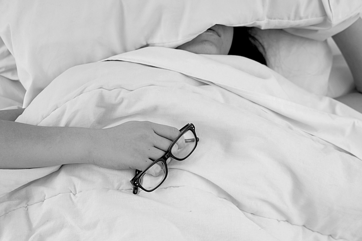Eine kranke / schlafende Frau, die im Bett liegt und das Kissen halb über ihr Gesicht gezogen hat und eine Brille in der Hand trägt